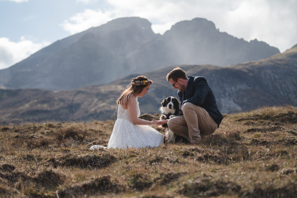 an elopement with a view across to Bla Bheinn an the Isle of Skye. The couple are holding paws with their collie dogs with the mountains behind them.e
