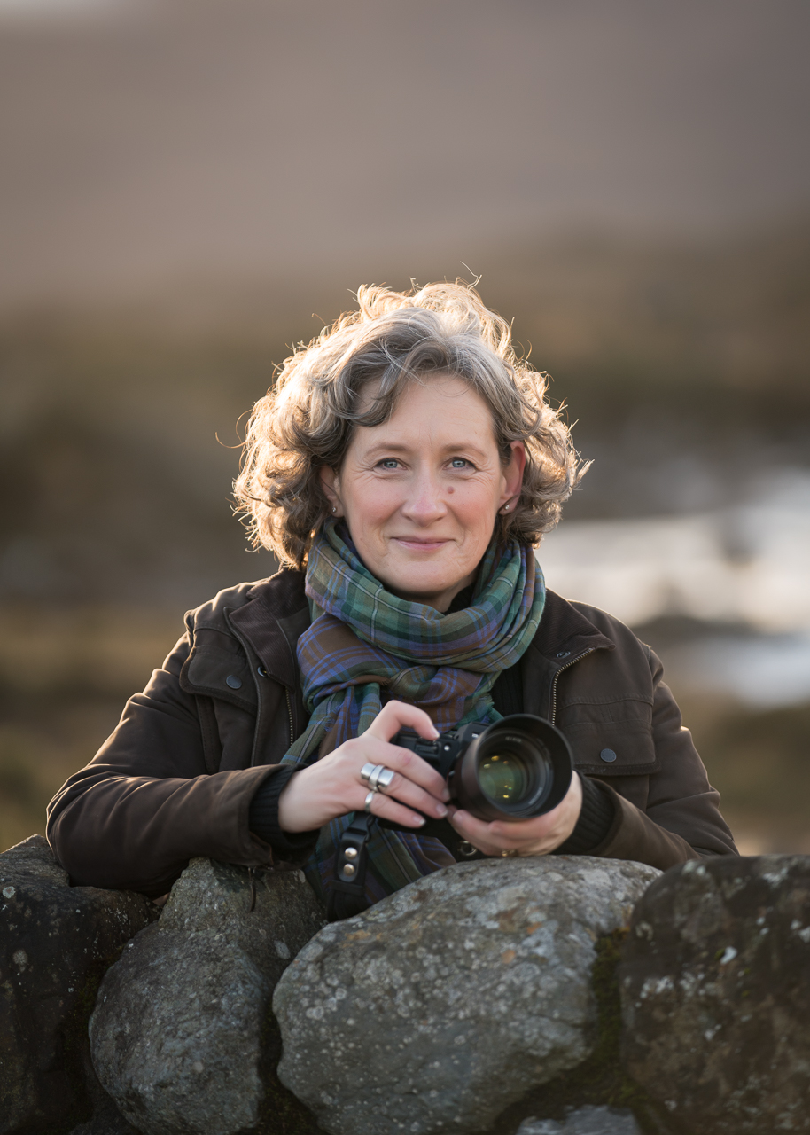 Picture of Penny Hardie, Isle of Skye Photographer, standing by a dry stone wall and holding a camera